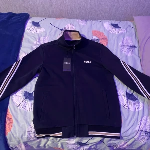  Mörkblå Hugo Boss dress - Snygg mörkblå dress från BOSS med dragkedja och vita ränder på ärmarna. Jackan har en hög krage och ribbade muddar vid ärmslut och nederkant. Perfekt för en stilren look.