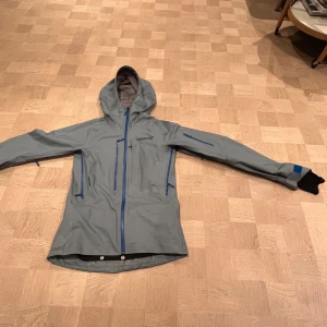 Grå Gore-Tex jacka från Norrøna - Säljer en grå Gore-Tex Lofoten jacka från Norrøna med huva och flera praktiska fickor med dragkedja. Jackan har en stilren design med blå detaljer och är perfekt för utomhusaktiviteter. Den är långärmad och tillverkad i ett slitstarkt material som skyddar mot väder och vind.