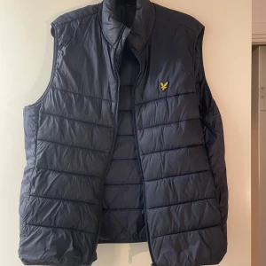 Mörkblå dunväst med broderad logga - Snygg mörkblå dunväst med en broderad logga på bröstet. Västen har en hög krage och är quiltad för extra värme. Perfekt för kyliga dagar.