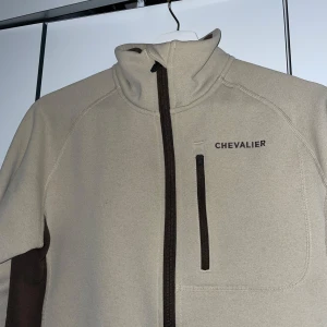 Beige fleecejacka från Chevalier - Säljer en beige fleecejacka från Chevalier med bruna detaljer. Jackan har en dragkedja framtill och en hög krage. Perfekt för kyliga dagar. Använts ett väldigt fåtal gånger .