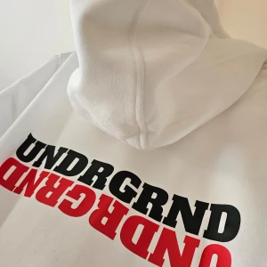 Vit hoodie med tryck från UNDRGRND - Snygg vit hoodie från UNDRGRND med tryck i svart och rött. Den har en känguruficka och justerbar huva. Perfekt för en avslappnad stil.