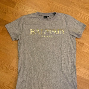  Balmain T-shirt - Snygg grå t-shirt från Balmain med guldfärgad logotyp på bröstet. Perfekt för en stilren och lyxig look. T-shirten har en normal passform och är kortärmad.