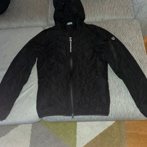 Svart quiltad jacka från EA7 Emporio Armani - Snygg svart quiltad jacka från EA7 Emporio Armani med huva och dragkedja. Perfekt för kyligare dagar med stilren design och logotypdetalj på ärmen.