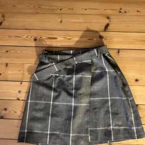 Grå rutig kjol med bälte - Snygg grå rutig kjol med ett matchande bälte. Perfekt för höst och vår, med en klassisk och stilren look. Kjolen har en normal passform och är gjord i ett bekvämt material. Den är idealisk för både vardag och festligare tillfällen.
