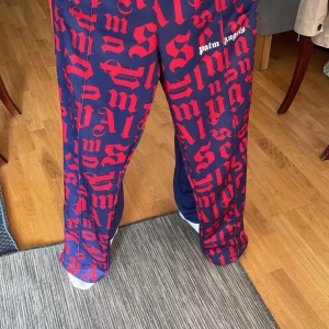 Blå och röda byxor från Palm Angels - Säljer ett par coola blå byxor med rött mönster från Palm Angels. Byxorna har en loose passform och är perfekta för en avslappnad stil. De har en elastisk midja och sidoremsor i rött. Perfekta för höst och vår! Digital kvitto finns också dem är L men passar även om man har m 