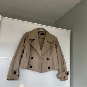 Beige kavaj från Zara - Säljer en snygg beige kavaj från Zara i bra skick. Den är croppad och har stora svarta knappar framtill. Perfekt för höst och vår, med en stilren look som passar både till vardags och fest. Stor krage och långa ärmar med knappar vid ärmsluten.