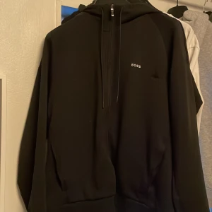 Svart hoodie från Hugo Boss - Säljer en stilren svart hoodie från Hugo Boss. Den har en dragkedja framtill och en klassisk huva med snören. Perfekt för både vardag och avslappnade tillfällen. Märket 'BOSS' är diskret tryckt på bröstet. Passar perfekt till höst och vinter.