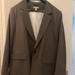 Brun kavaj - Snygg brun kavaj i klassisk stil. Perfekt för både formella och semi-formella tillfällen. Kavajen har en enkel knäppning fram och två fickor. Passar bra till höst- och vintergarderoben.