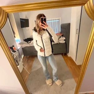 Vit pufferjacka med svarta detaljer - Säljer en snygg vit skidjacka med svarta detaljer och dragkedja. Jackan har en huva och är perfekt för fjällen. Den är i mycket bra skick och håller värmen. Storleken är dam S och den har en normal passform.