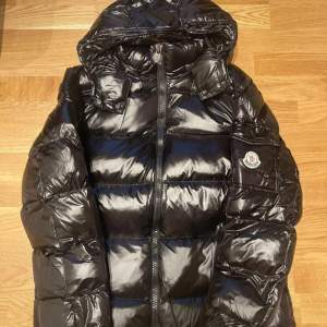 Säljer en snygg svart dunjacka från Moncler i mycket bra skick. Jackan har en glansig finish och en praktisk huva. Den är perfekt för kalla vinterdagar och har en dragkedja framtill. Moncler-loggan sitter på ärmen. Perfekt för dig som vill hålla stilen i vinter! ❄️