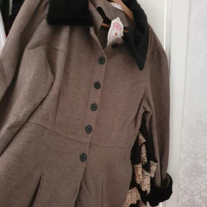 Brun kappa från Tesettür Dünyası - Säljer en elegant brun kappa från Tesettür Dünyası i storlek 46. Den har en klassisk design med svarta knappar och en mjuk krage i svart pälsimitation. Perfekt för höst och vinter med sina långa ärmar och stilrena look. Kappan är i mycket bra skick och redo för nya äventyr!