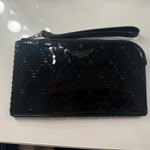 Svart glansig plånbok med dragkedja  - Snygg svart plånbok i glansigt material med quiltat mönster och en cool metallisk detalj på framsidan. Den har en praktisk dragkedja och en handledsrem för enkel användning. Perfekt för att hålla ordning på kort och kontanter.