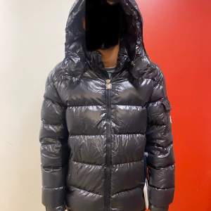 Säljer en snygg svart dunjacka från Moncler i mycket bra skick. Jackan har en glansig finish och är perfekt för kalla vinterdagar. Den har en praktisk huva och dragkedja framtill. Moncler-loggan sitter på ärmen. Perfekt för dig som vill hålla stilen i vinter! 🖤