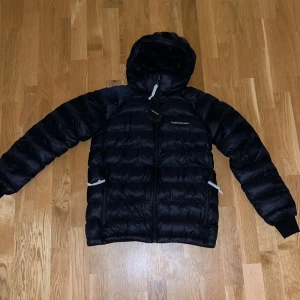 Svart dunjacka från Peak Performance - Säljer en svart dunjacka från Peak Performance som är väldigt eftertraktad. Storlek 170 JuniorStorlek. Jackan är i nytt skick med etikett kvar. Den har en praktisk huva och dragkedja framtill. Perfekt för kalla vinterdagar! 🖤Säljes Pga fel köp av storlek. Helt oanvänd. Jackan är i Unisex passform. 