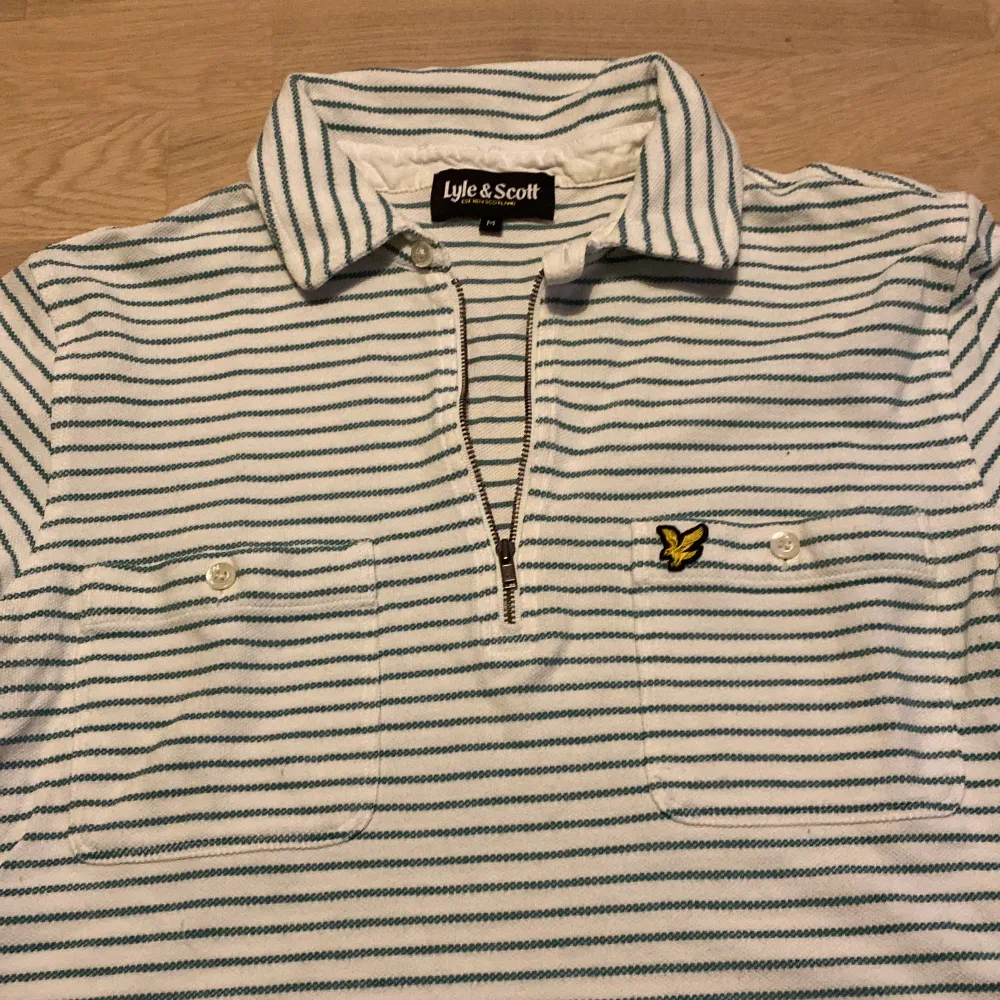 Säljer en snygg randig pikétröja från Lyle & Scott i storlek M. Tröjan är vit med gröna ränder och har en klassisk krage med dragkedja. Den har korta ärmar och två bröstfickor med knappar. Perfekt för en avslappnad stil!. Tröjor & Koftor.