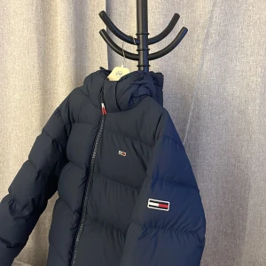 Mörkblå dunjacka med huva - Säljer en snygg och varm mörkblå dunjacka från Tommy Hilfiger. Jackan har en praktisk huva och dragkedja framtill. Perfekt för kalla vinterdagar.  Skick 9/10 då den är helt oanvändbar. Ord pris 1750kr