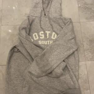 Grå hoodie med Boston-tryck - Säljer en grå hoodie med texten 'Boston South' på bröstet. Den är supermjuk och perfekt för kyliga dagar. Har en klassisk känguruficka och en justerbar huva. Perfekt för en avslappnad stil eller mysiga hemmakvällar. 🐨
