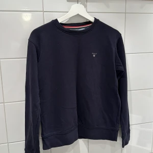 Mörkblå tröja från GANT - Säljer en stilren mörkblå tröja från GANT i mycket bra skick. Den har en klassisk design med rund hals och långa ärmar. Perfekt för både vardag och lite finare tillfällen. Passar bra till jeans eller chinos! Stl 170.