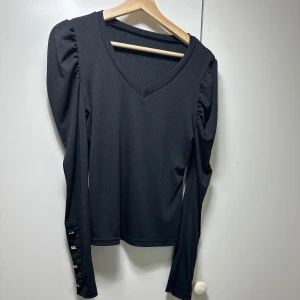 Svart ribbad topp med knappar - Säljer en stilren svart ribbad topp med v-ringning och långa ärmar. Toppen har snygga knappar på ärmsluten som ger en extra touch. Perfekt för både vardag och fest!