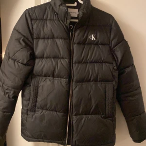 Svart pufferjacka från Calvin Klein - Säljer en snygg svart pufferjacka från Calvin Klein i mycket bra skick. Jackan har en dragkedja framtill och är quiltad för extra värme. Perfekt för höst och vinter! Den har en stilren design med CK-logga på bröstet. Priset är ej hugget i sten 