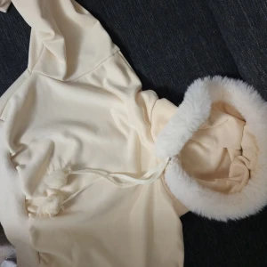 Beige hoodie med fluffig huva - Mysig beige hoodie med fluffig vit kant runt huvan. Perfekt för kalla dagar med sina långa ärmar och mjuka material. Har snören med bollar för justering av huvan. Superfin och bekväm för både vardag och mysiga hemmakvällar.