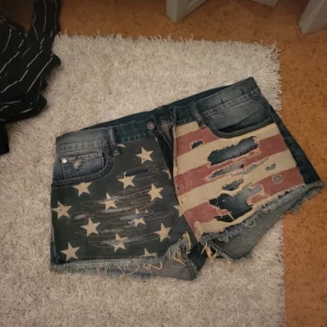 Raulph Lauren cut off shorts  - Säljer ett par unika jeansshorts med coolt stjärn- och randmönster. Ena sidan har vita stjärnor på blå bakgrund och den andra sidan har slitna ränder i rött och beige. Perfekta för sommarens festivaler eller en dag på stranden. De har en lågmidjad passform och fransiga kanter för en avslappnad look, dessa shorts är från Raulph Lauren.