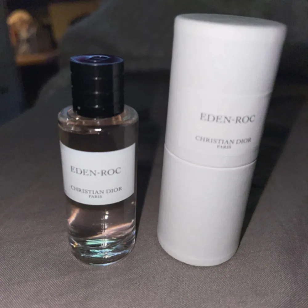 Säljer en elegant flaska av Eden-Roc parfym från Christian Dior. Flaskan är genomskinlig med en svart kork och har en stilren vit etikett. Parfymen kommer i en vit cylinderformad förpackning. Perfekt för den som älskar lyxiga dofter!. Perfume.