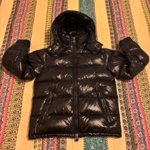 Moncler Maya Puffer Jacka - Säljer en snygg svart pufferjacka från Moncler i ny skick. Jackan har en glansig finish och är perfekt för kalla vinterdagar. Den har en praktisk huva och dragkedja framtill. Perfekt för dig som vill hålla stilen i vinter! ❄️