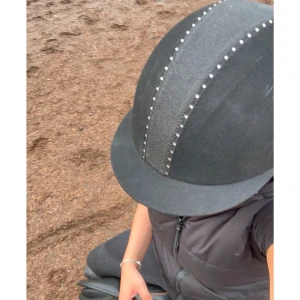 Svart ridhjälm med strassdetaljer - Snygg svart ridhjälm med glittriga strassdetaljer längs mitten. Perfekt för ridning och ger en stilren look samtidigt som den skyddar. Hjälmen har en slät yta och en klassisk form som passar de flesta. Perfekt för både träning och tävling! Den är lite smutsig på bilden men gör rent den innan den blir fraktad.