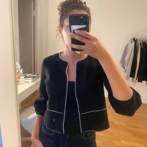 Svart kofta med vita detaljer - Säljer en stilren svart kofta med vita kanter. Den har trekvartsärmar och en elegant passform. Perfekt för både vardag och fest. Passar bra till jeans eller klänning.