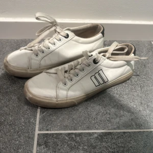 Vita sneakers med svart detalj - Säljer ett par vita sneakers i bra skick med svart detalj på sidan, köpta i Spanien . De har snörning och en klassisk design som passar perfekt till vardags. Sulan är lite smutsig men annars är de i fint skick. Perfekta för både vår och sommar!