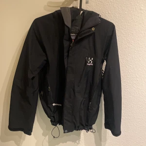 Svart vindjacka från Haglöfs - Säljer en svart vindjacka från Haglöfs i bra skick. Jackan har en praktisk huva och dragkedja framtill. Perfekt för höst- och vårväder med sin slitstarka design och justerbara midja. Passar både till vardags och utomhusaktiviteter.