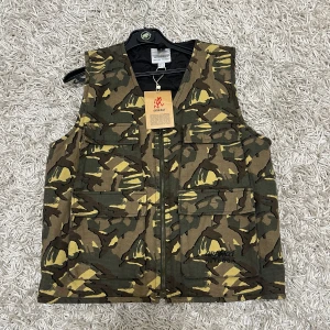 Gramicci väst - Säljer en helt ny camouflageväst från Gramicci i storlek XL. Passar även längden runt 175-180cm. Västen har ett klassiskt camouflagemönster i gröna och bruna toner med dragkedja framtill. Perfekt för utomhusaktiviteter eller som en stilren detalj i garderoben.