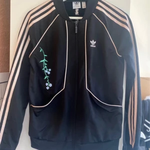 Svart Adidas tröja med blommor - Säljer en snygg svart Adidas tröja med rosa ränder och blommiga broderier. Tröja  har dragkedja och långa ärmar, perfekt för vår och höst. På baksidan finns texten 'Love all, trust a few, do wrong to none'.