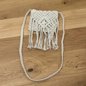 Vit axelväska i makramé - Säljer en superfin vit axelväska i makramé med fransar. Den har ett snyggt flätat mönster och en praktisk dragkedja upptill. Perfekt för sommarens festivaler eller en dag på stan. Väskan är i mycket bra skick och redo för nya äventyr!