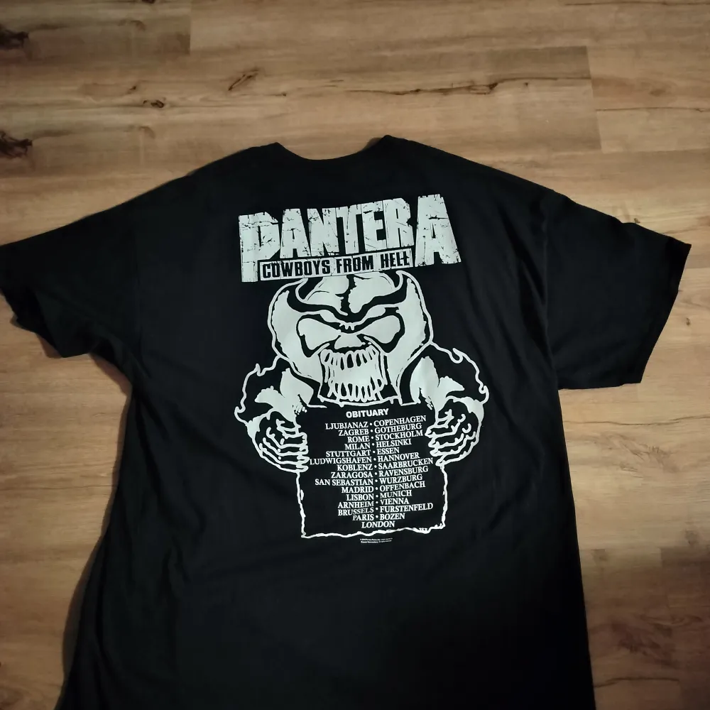 Säljer en svart T-shirt med Pantera-tryck 'Cowboys from Hell'. Framsidan har en cool design med en dödskalle i cowboyhatt och blodiga detaljer. Baksidan listar turnéstäder med en stor dödskalle. Perfekt för rockfans! 🖤. T-shirts.