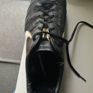 Svarta fotbollsskor från Nike - Säljer ett par svarta fotbollsskor från Nike i bra skick. Skorna har snörning och är perfekta för fotbollsträning. De har en klassisk design med vita detaljer och dobbar för bra grepp på planen.