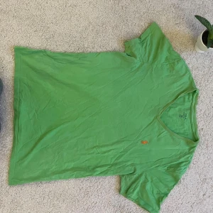 Grön t-shirt från Ralph Lauren - Säljer en grön t-shirt från Ralph Lauren med en broderad logga i orange på bröstet. T-shirten har en v-ringad hals och korta ärmar. Perfekt för en avslappnad stil.