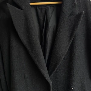 Mörkblå kappa i ullblanding  - Mörkblå kappa i ullblanding i storlek 34. Köpt på Åhléns för några år sedan. I gott skick. Dubbelknäppning framtill. Något oversize i passformen.