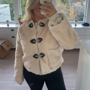 Beige pälsjacka med knäppning - Säljer en mysig beige pälsjacka med unika knäppdetaljer framtill. Jackan har långa ärmar och en mjuk, fluffig yta som är perfekt för kyligare dagar. Perfekt för dig som vill ha en stilren och varm look.