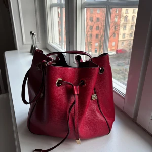 Röd axelväska med snörning - Snygg röd axelväska i läder med praktisk snörning och guldfärgade detaljer. Perfekt för att ge en färgklick till din outfit. Väskan har en rymlig design