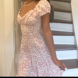 Blommig sommarklänning - Charmig kortklänning med blommigt mönster i rosa och vitt. Klänningen har en söt v-ringning och korta ärmar. Perfekt för varma sommardagar.