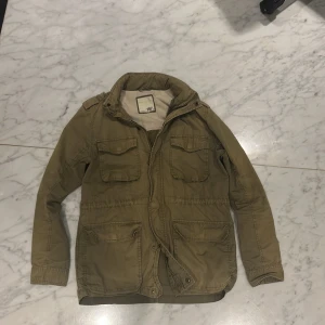 Mörkgrön fieldjacka från 157 - Säljer en mörkgrön fieldjacka från 157 med inbyggd huva och dragkedja. Jackan har flera praktiska fickor och är perfekt för allt väder. Den har långa ärmar och en stilren design som passar till det mesta. Perfekt för dig som vill ha en funktionell och snygg jacka. Pris lätt diskuterbart vid snabb affär 