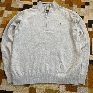 Grå tröja med dragkedja från Gant - Säljer en stilren grå tröja från Gant med halv dragkedja och broderad logga på bröstet. Tröjan har långa ärmar och ribbade muddar vid ärmslut och nederkant. 