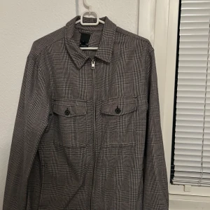 Grå rutig overshirt - Snygg grå rutig overshirt med dragkedja och två bröstfickor med knappar. Perfekt för lager-på-lager-stil. Den har en klassisk krage och långa ärmar, vilket gör den idealisk för både höst och vår. Använts några gånger storlek S men skulle säga att den passar bättre för M. Dm för fler bilder och pris kan även diskuteras. 