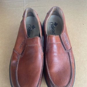 Bruna loafers från Rieker - Snygga bruna loafers från Rieker i skinn. De har en klassisk design med bekväm passform och robust sula. Perfekta för en stilren look.