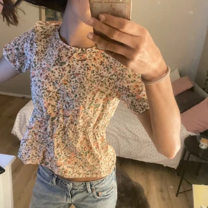 Blommig söt blus till sommaren  - Söt blommig blus med korta ärmar och rund hals. Blusen har ett färgglatt mönster i rosa, grönt och orange på en vit bakgrund. Den har en knappdetalj i nacken för enkel på- och avtagning.