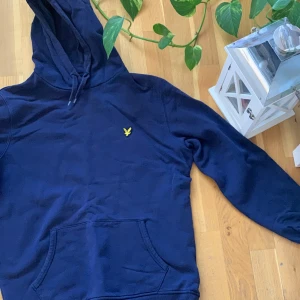 Mörkblå hoodie från Lyle & Scott - Säljer en snygg mörkblå hoodie från Lyle & Scott med en gul logga på bröstet. Tröjan har en klassisk känguruficka och justerbar huva med dragsko. Perfekt för en avslappnad stil. Aldrig använd. Nypris 1500 kronor. 