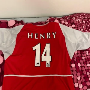 Röd Arsenal fotbollströja med Henry 14 - Säljer en klassisk röd Arsenal fotbollströja från Nike med Henrys namn och nummer 14 på ryggen. Tröjan har vita ärmar och klubbens emblem på bröstet. Perfekt för fans av Arsenal och fotbollshistoria. Tröjan är köpt för över 20 år sedan i just london och är size XL men passar minst lika bra på L.