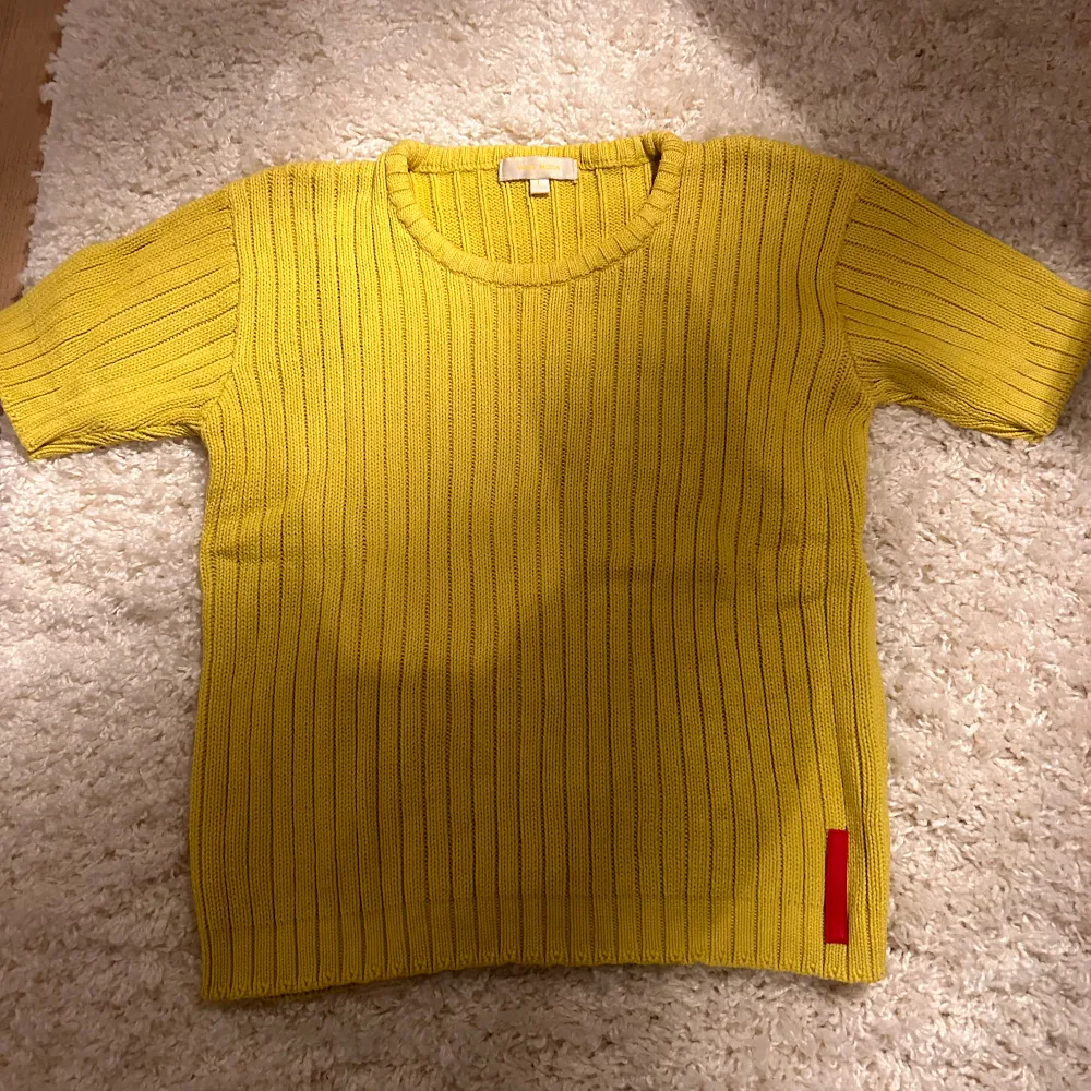 Säljer en pippigul stickad tröja från Vero Moda. Stl L men den är mer som M. Tröjan har korta ärmar och ett ribbat mönster som ger en stilren look. Perfekt för att lysa upp garderoben med en färgklick. Detalj i rött vid nederkanten ger en extra touch.. T-paidat.